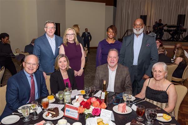 A group of eight well-dressed adults pose for a photo at a formal dinner event. Four individuals are standing behind a round table while four others are seated at the table. The table is set with glasses, plates of cake, salads, rolls, and bottles of wine and water. The background shows a large room